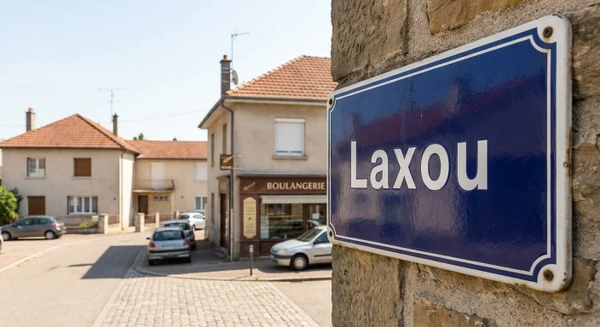 Plan rapproch&eacute; d'une plaque de rue ou d'une signal&eacute;tique urbaine indiquant "Laxou" dans un cadre de petite commune en agglom&eacute;ration, avec en arri&egrave;re-plan flou un environnement urbain typique de la banlieue nanc&eacute;ienne (petits commerces, rue calme, b&acirc;timents r&eacute;sidentiels). Photo de jour en conditions d'&eacute;clairage naturel, perspective r&eacute;aliste et professionnelle.