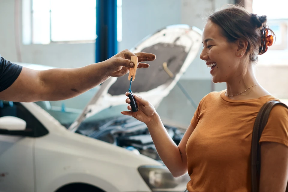 Atelier automobile moderne avec mécanicien en combinaison bleue consultant documents jaunes avec cliente en chemisier beige près d'un véhicule noir au capot ouvert